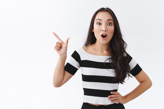 Geamuseerd vroeg zich af knappe oost-aziatische brunette vrouw in gestreepte t-shirt open mond hijgend verbaasd en kijk camera geïntrigeerd wijzend linker bovenhoek als interessante promo bespreken