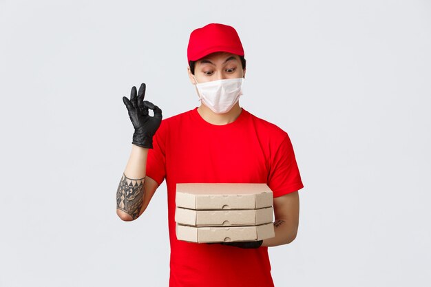 Geamuseerde Aziatische bezorger rood uniform, pet en t-shirt, met beschermende handschoenen en medisch masker voor de veiligheid van de klant, als eten bezorgen bij de deur, pizza brengen, goed teken tonen en naar smakelijke pizza staren