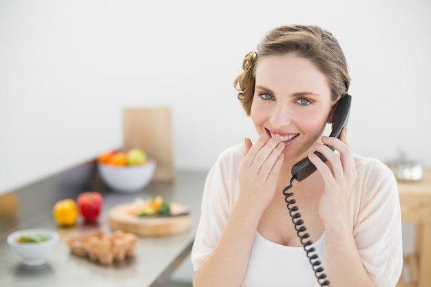 Geamuseerde mooie vrouwenzitting in haar keuken die met een telefoon telefoneert