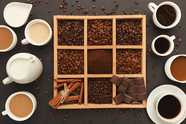 Geassorteerde gemalen koffie en geroosterde bonen, kruiden en chocolade in houten vierkant frame en verschillende koffiekopjes op grijze getextureerde leisteen, bovenaanzicht, kopieerruimte. Sjabloonontwerp voor het voorkomen van café