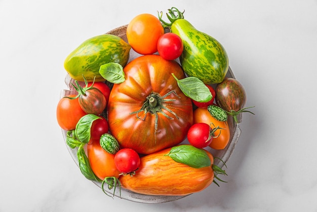 Geassorteerde kleurrijke biologische tomaten met minikomkommers en basilicum in een bord op witte tafel Bovenaanzicht