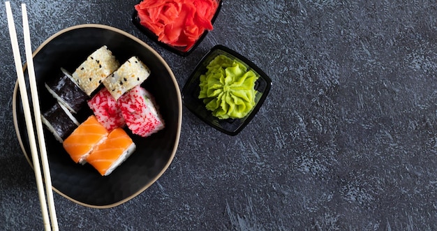 Geassorteerde sushibroodjes liggen op een zwarte plaat met wasabi-gepekelde gember en bamboestokjes op een donkere achtergrond met een kopieerruimte aan de rechterkant Bovenaanzicht platte lay-out horizontaal