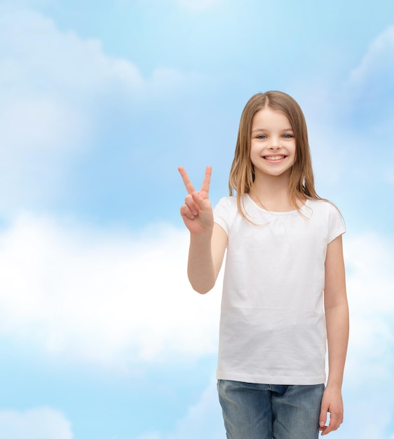 gebaar en gelukkig mensenconcept - glimlachend meisje in wit leeg t-shirt dat vredesgebaar met vingers toont