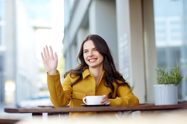 gebaar, levensstijl, drankjes en mensenconcept - gelukkige jonge vrouw of tienermeisje met kopje cacao drinken en zwaaiende hand op caféterras in de stad