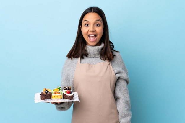 Gebakjechef-kok die een grote cake over blauwe muur met verraste gelaatsuitdrukking houden.