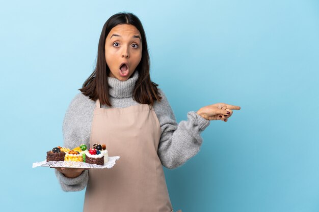 Gebakjechef-kok die een grote cake over geïsoleerd verrast blauw houden en kant richten