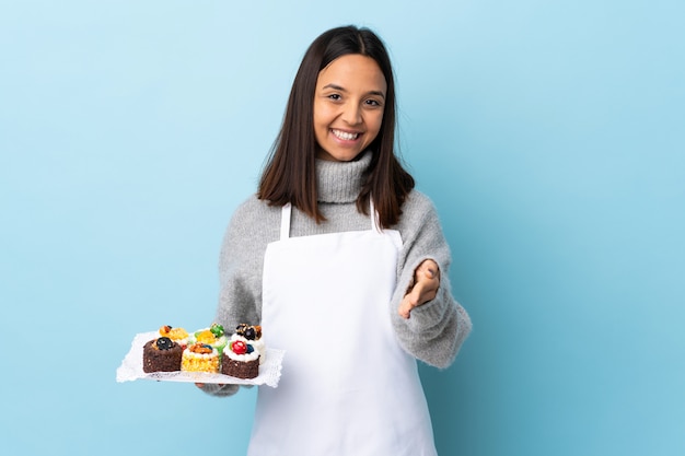 Gebakjechef-kok die een grote cake over geïsoleerde blauwe achtergrond houden die handen schudden voor het sluiten van een goede deal