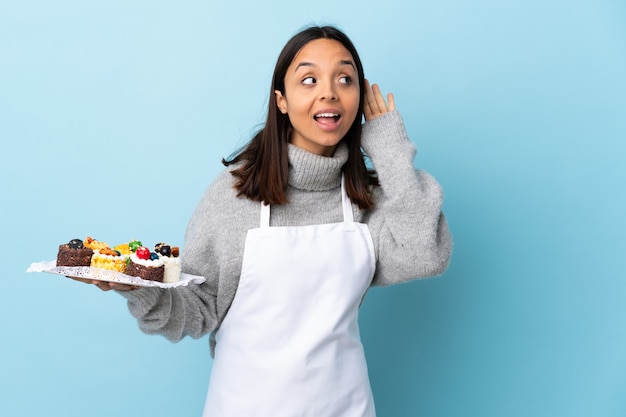 Gebakjechef-kok die een grote cake over geïsoleerde blauwe achtergrond houden luisterend aan iets door hand op het oor te zetten