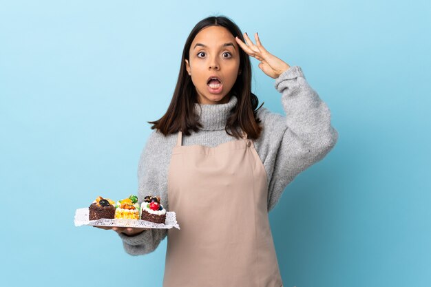 Gebakjechef-kok die een grote cake over geïsoleerde blauwe achtergrond met verrassingsuitdrukking houden