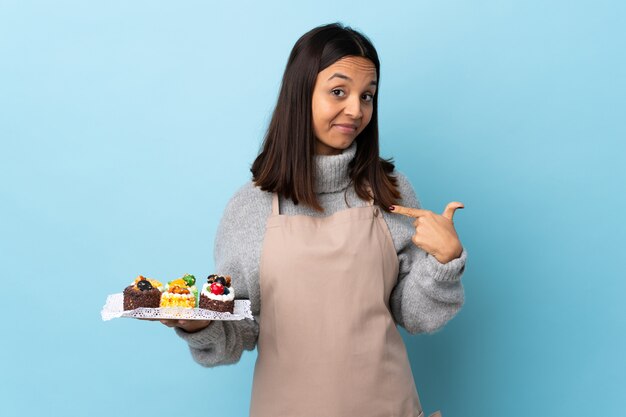 Gebakjechef-kok die een grote cake over geïsoleerde blauwe muur trots en zelfvoldaan houden
