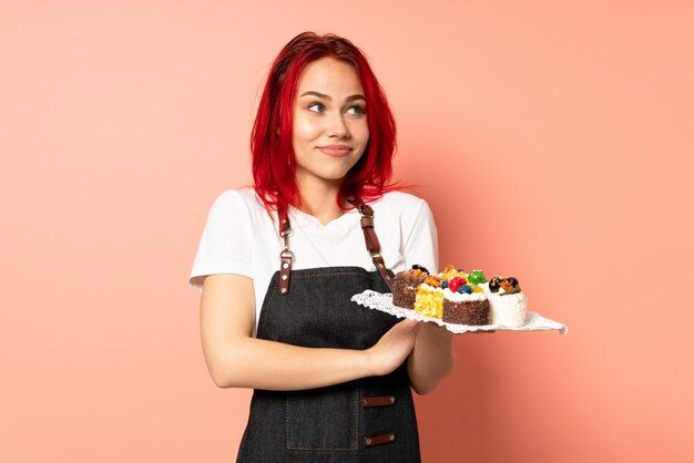 Gebakjechef-kok die muffins houden die op roze muur worden geïsoleerd die twijfelsgebaar maken terwijl het opheffen van de schouders