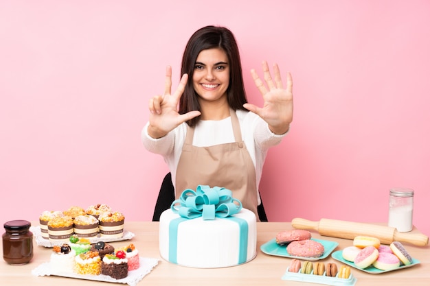 Gebakjechef-kok met een grote cake in een lijst over geïsoleerde roze muur die acht met vingers tellen