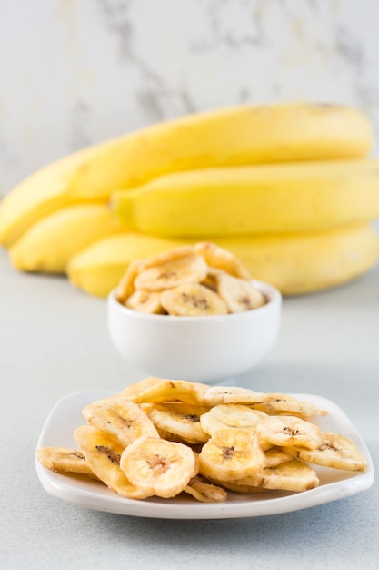 Gebakken bananenchips in een witte kom en schotel en een tros bananen op tafel