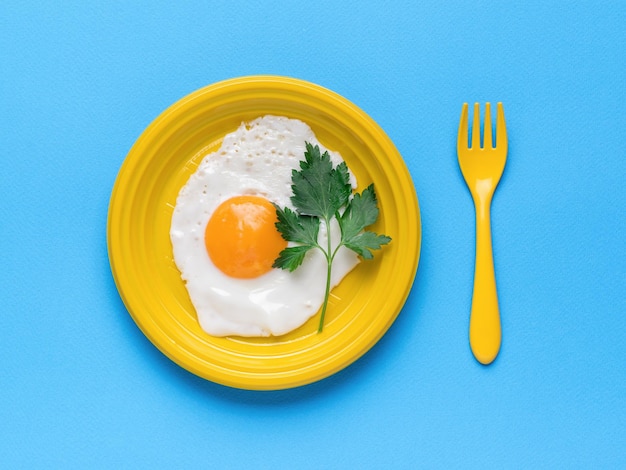 Gebakken ei en een takje peterselie op een gele plastic plaat en een vork op een blauwe achtergrond Een populair ontbijt Minimaal concept Een populair ontbijt