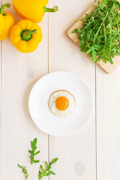 Gebakken ei in een bord met rucola en paprika op een witte houten tafel