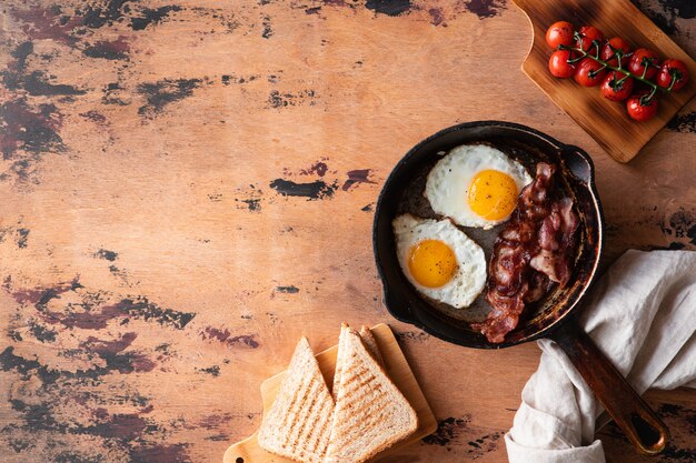 Gebakken eieren in een koekenpan met zongedroogde cherrytomaatjes, spek en geroosterd brood voor het ontbijt op een rustiek hout