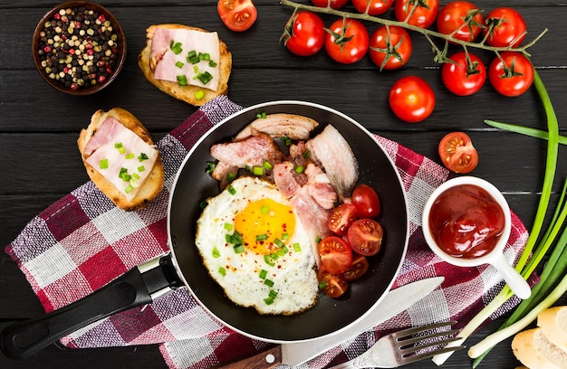 Gebakken eieren met ham en tomaten in pan op zwarte houten achtergrond top view