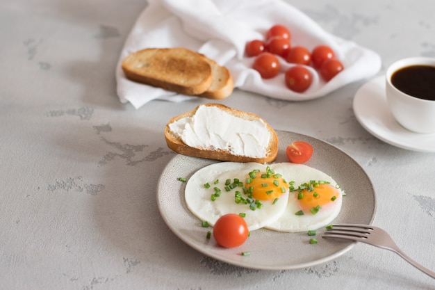 Gebakken eieren met tomaten en groene uien op een grijze plaat, toast en zwarte koffie op een lichte achtergrond