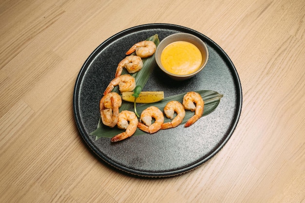 Gebakken garnalen op een groen blad met een schijfje sinaasappel en gele saus op een zwarte plaat op een houten tafel achtergrondxD