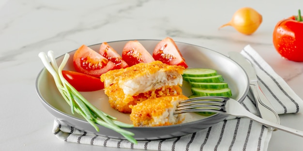 Gebakken gepaneerde visfilets met komkommer mier tomaten geserveerd op een bord close-up.