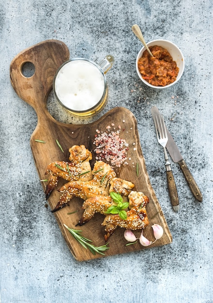 Gebakken kippenvleugels op rustieke portie bord, pittige tomatensaus, kruiden en mok licht bier over zwarte houten tafel