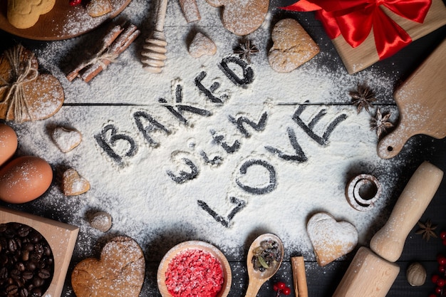 Foto gebakken met liefde geschreven op bloem. peperkoek hartvormige koekjes, kruiden, koffiebonen en bakbenodigdheden op zwarte houten achtergrond