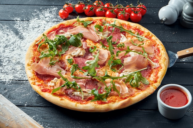 Gebakken pizza met salami, prosciutto en kip met rode saus en gesmolten kaas op een zwarte houten tafel in een compositie met ingrediënten. Bovenaanzicht