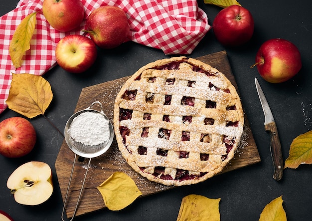 Gebakken ronde traditionele appeltaart op bruin houten plank en verse rode appels bovenaan