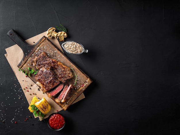 Gebakken steaks met kruiden en specerijen op houten bord perkament gegrilde knoflook maïs twee sauzen op zwarte achtergrond close-up bovenaanzicht