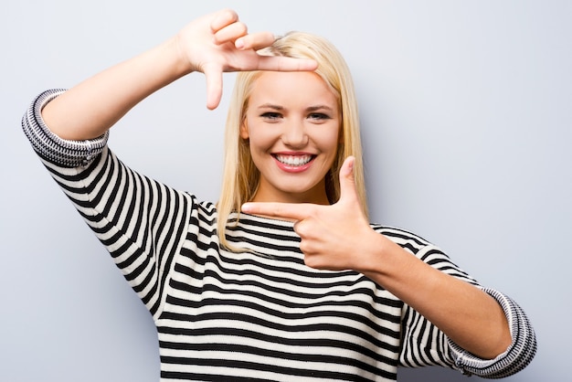 Gebaren vinger frame. Mooie jonge blonde haarvrouw die naar de camera kijkt en een vingerframe gebaart terwijl ze tegen een grijze achtergrond staat