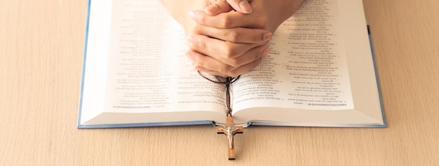 Gebedende mannelijke hand die het kruis vasthoudt op het heilige bijbelboek aan een houten tafel