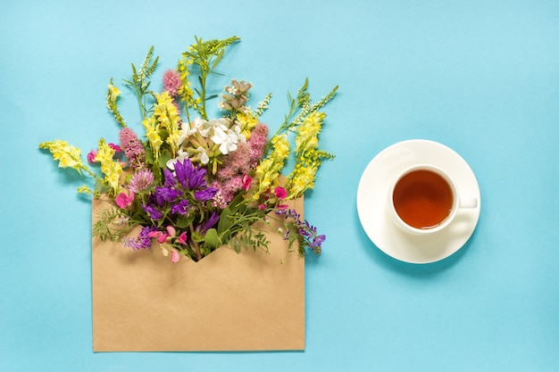 Gebieds kleurrijke bloemen in ambachtenvelop en kop thee op blauw