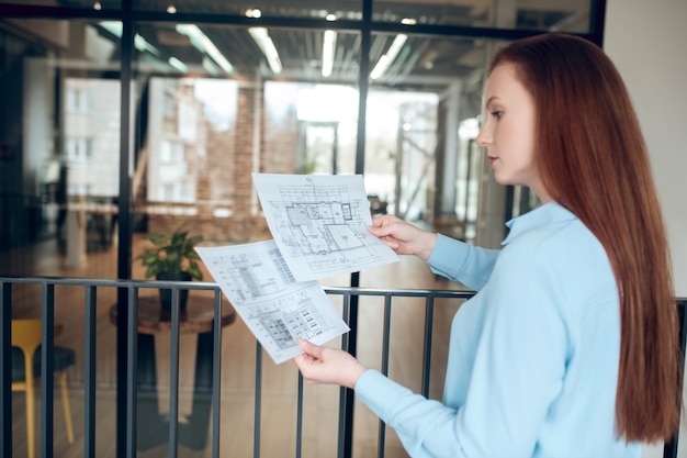 Gebouw functie. Slimme jonge volwassen knappe vrouw met lang rood haar die nadenkt over een bouwplan dat binnenshuis voor een glazen wand staat