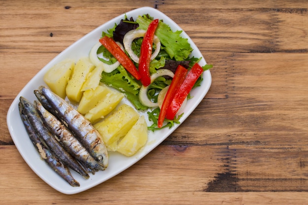 Gebraden sardines met gekookte aardappel en salade op witte plaat op bruin hout