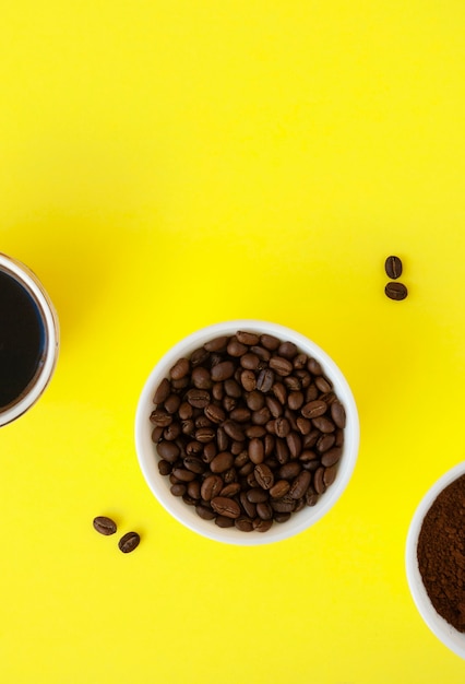 Gebrande koffiebonen, gemalen koffie en gebrouwen zwarte koffie in witte kommen op een gele ondergrond.