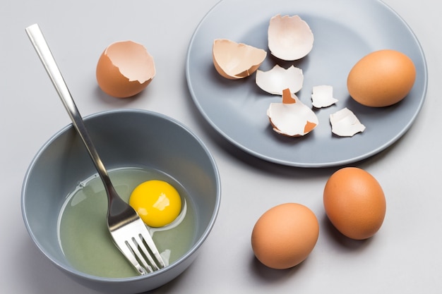 Gebroken ei en vork in grijze kom. Kippenshells op grijze plaat. Twee bruine eieren op tafel. Grijze achtergrond. Bovenaanzicht