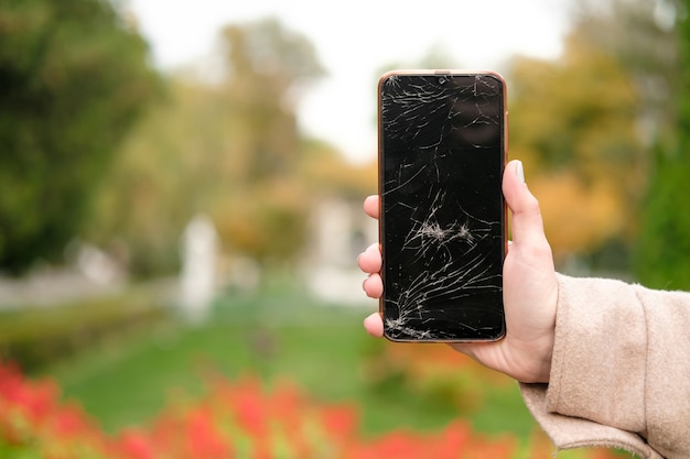 Gebroken glas op het telefoonscherm. Gebroken telefoon in de hand van een vrouw. Achtergrond van planten in het park. Onscherpe achtergrond