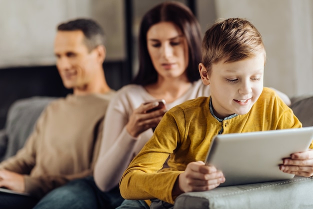 Gebruikelijk tijdverdrijf. Aangename kleine jongen die op de armleuning van de bank leunt en op de tablet speelt terwijl zijn ouders bezig zijn met hun eigen apparaten