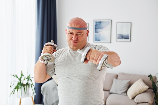 Gebruikt dumbbells Grappige man met overgewicht in vrijetijdskleding is binnenshuis thuis