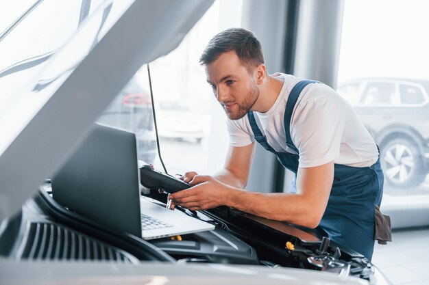 Gebruikt laptop Jonge man in wit overhemd en blauw uniform repareert auto