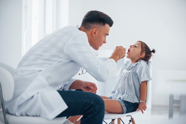 Gebruikt tonsillen om de keel te controleren. Jonge kinderarts werkt met kleine vrouwelijke bezoeker in de kliniek.