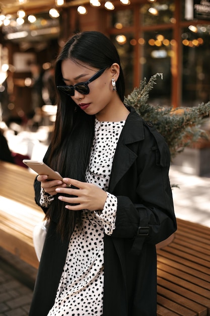 Gebruinde Aziatische vrouw houdt telefoon Brunette jonge dame in zonnebril zwarte trenchcoat en jurk buiten berichten