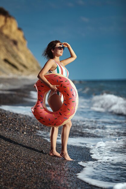 Gebruinde jonge vrouw in een zwempak en zonnebril die op een strand staat met een opblaasbare ring