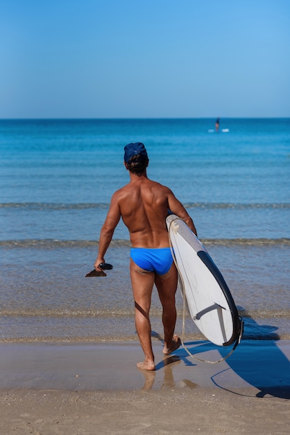 Gebruinde man op het strand met een surfplank en riem in zijn handen gaat het water in.