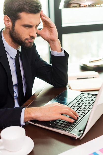 Geconcentreerd op het werk. Geconcentreerde jonge man in pak die hoofd met de hand aanraakt en naar laptop kijkt terwijl hij op zijn werkplek zit