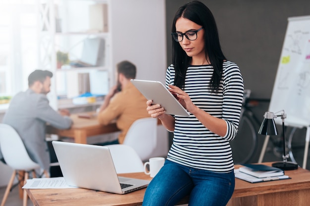 Geconcentreerd op het werk. Zelfverzekerde jonge vrouw die aan digitale tablet werkt terwijl haar collega's op de achtergrond werken