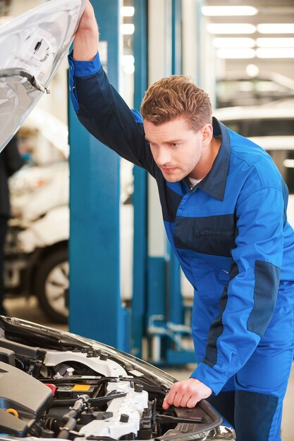 Geconcentreerd op probleem. Geconcentreerde jonge man in uniform die auto onderzoekt terwijl hij in de werkplaats staat