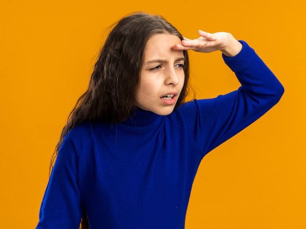 Geconcentreerd tienermeisje met loensende ogen die de hand op het voorhoofd houden en naar de zijkant kijken in de verte geïsoleerd op een oranje muur