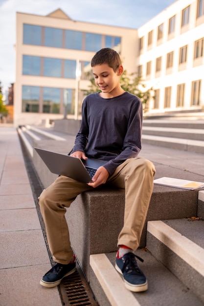 Foto geconcentreerde adolescente jongen die op de veranda van een gebouw zit en op de laptop typt