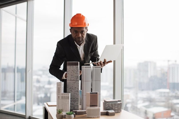 Geconcentreerde afrikaanse inspecteur die laptop gebruikt die wolkenkrabbermodel inspecteert in architectonisch modern kantoor